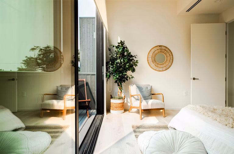 Modern bedroom with a wooden chair, potted plant, round mirror on the wall, open door, and sunlight coming through a large glass window.